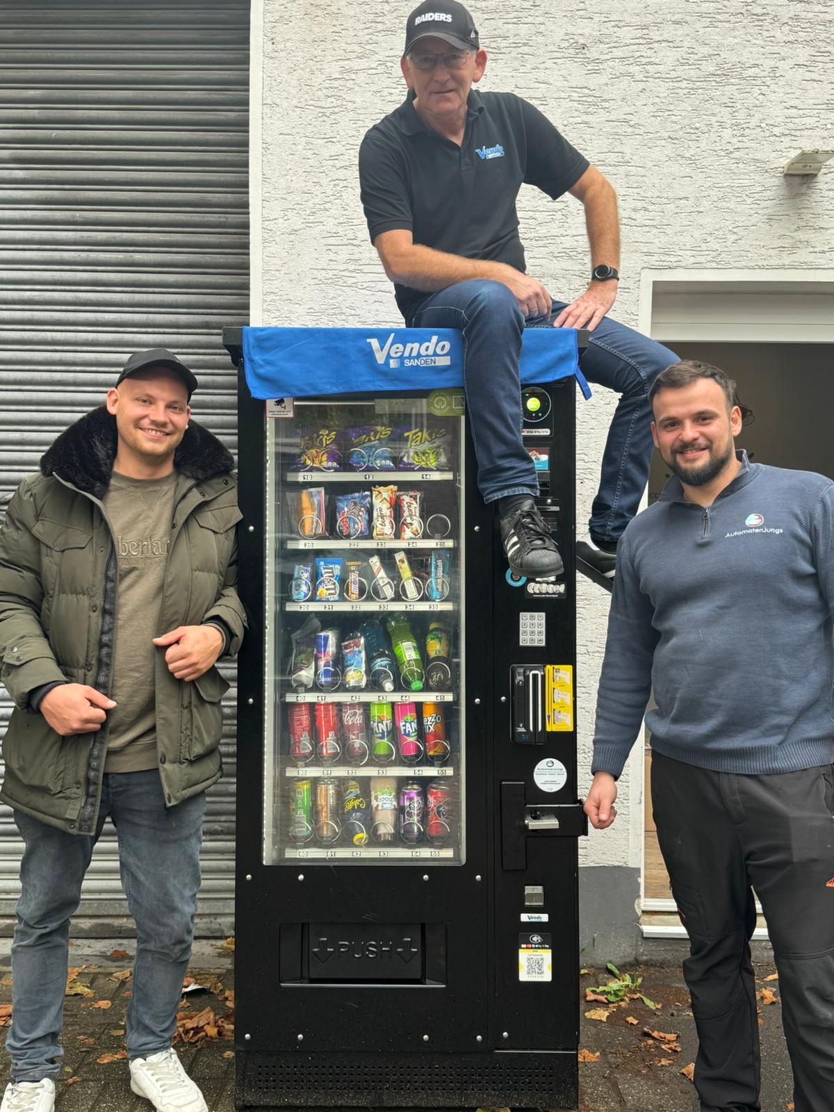 Drei Männer neben einem Vendo Snack- und Getränkeautomaten vor Gebäude